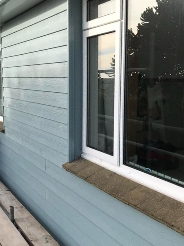 A white-framed window on a light blue wooden exterior wall, with brick trim below the window and trees reflected in the glass.