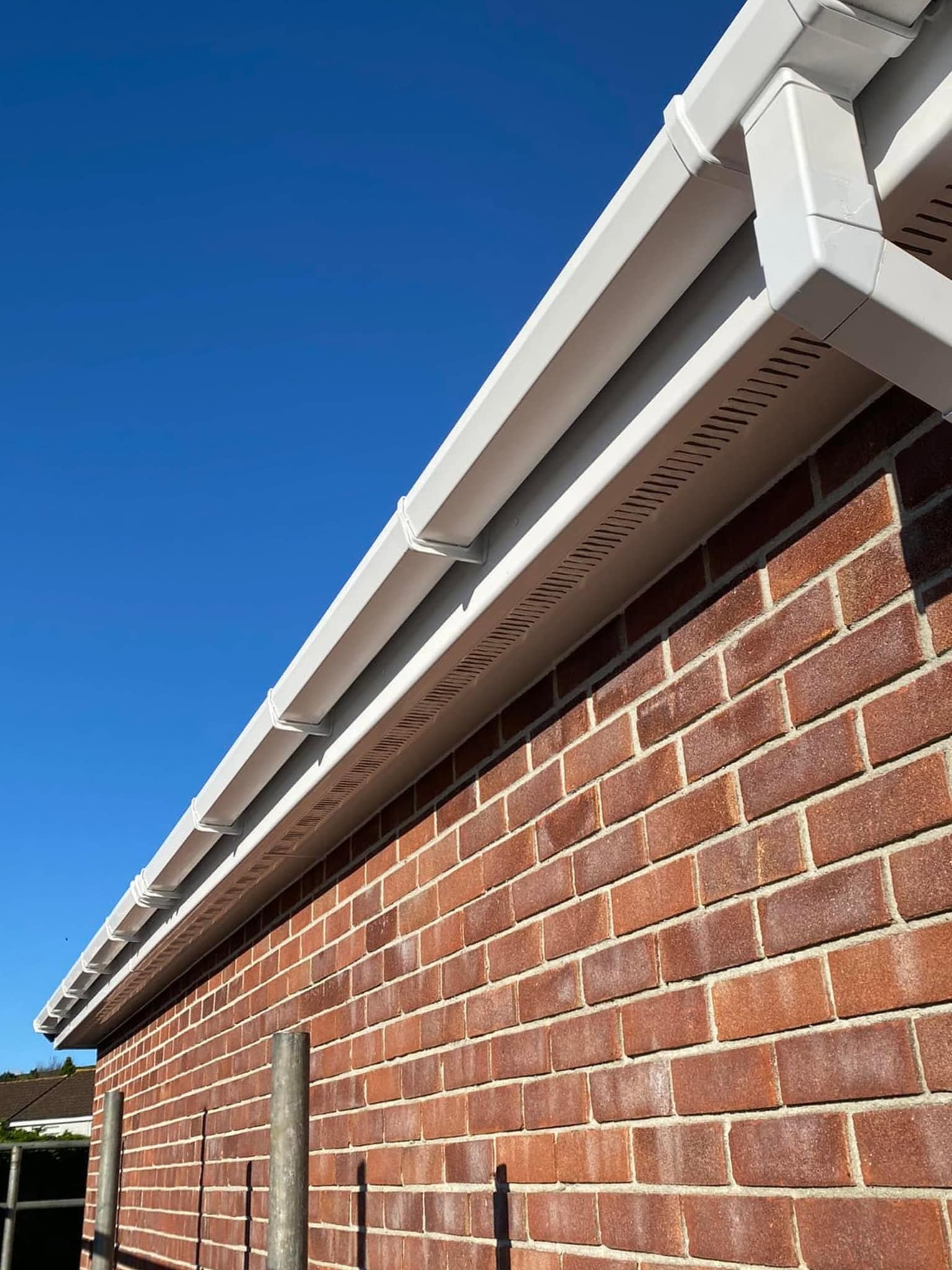 Close-up view of a red brick wall with white plastic guttering and soffit under a clear blue sky, shown from a low angle.
