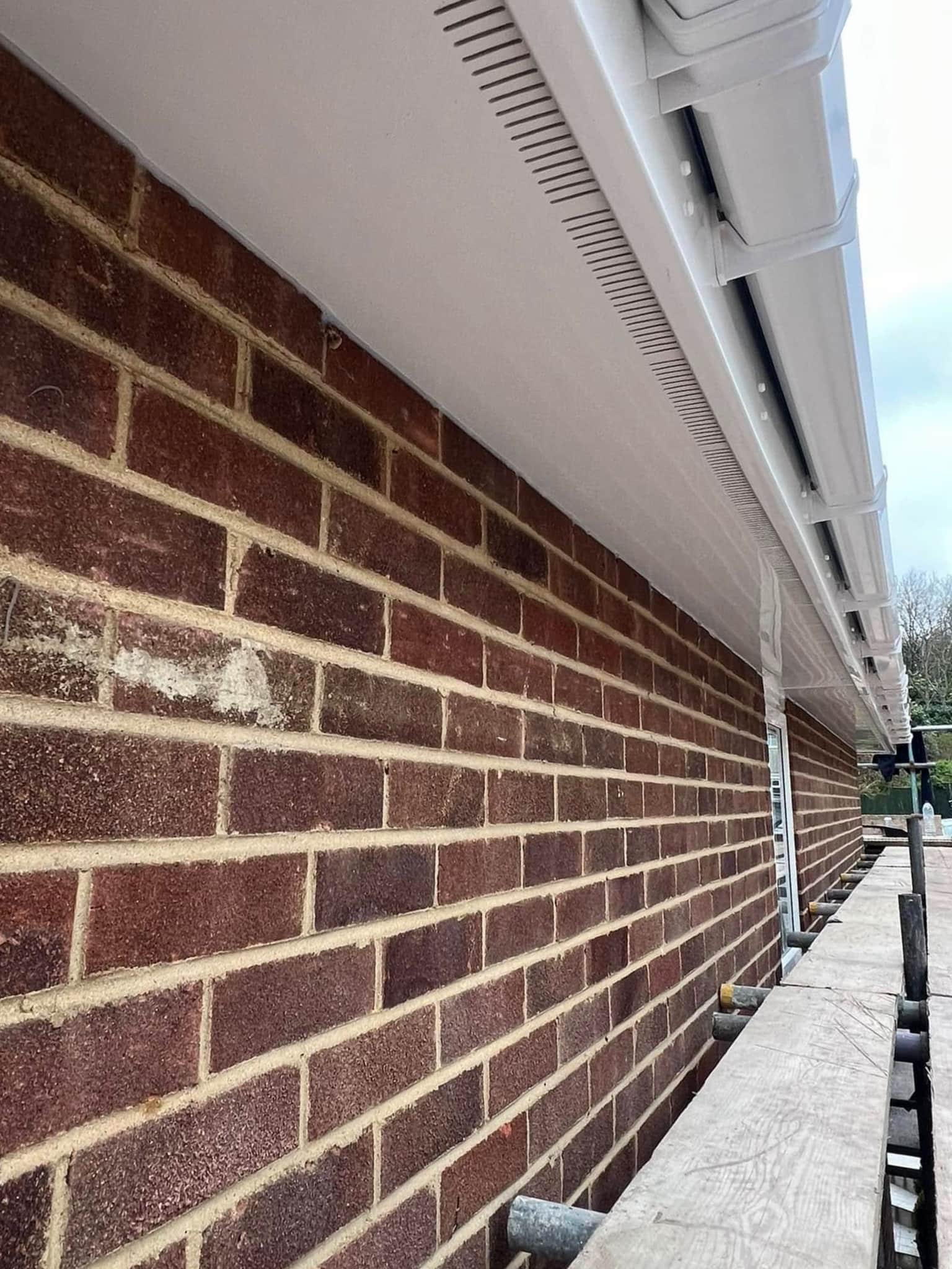 Close-up of a brick wall with newly installed white uPVC soffits and guttering along the roof edge, viewed from a scaffolding platform running parallel to the wall.