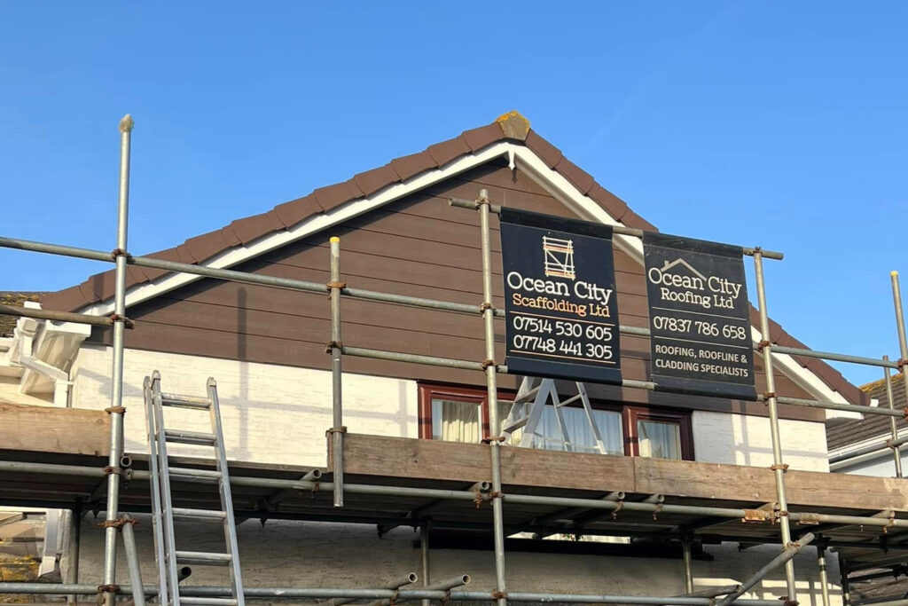 Scaffolding surrounds a house with two business signs: Ocean City Scaffolding Ltd and Ocean City Roofing Ltd. A ladder leans against the scaffold under a clear blue sky.