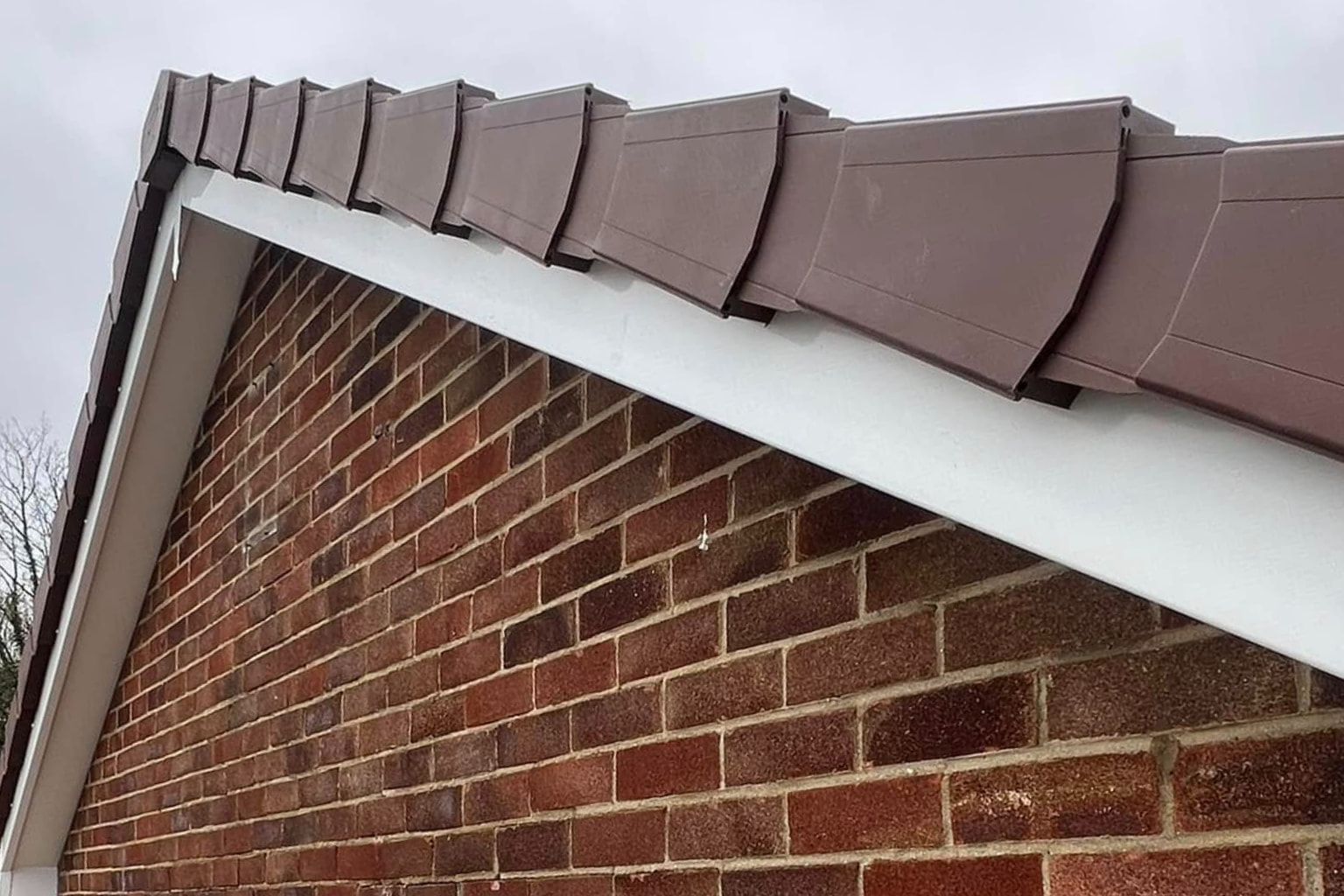 A close-up of a gable roof with brown roof tiles and white trim on a red brick building, under a cloudy gray sky.