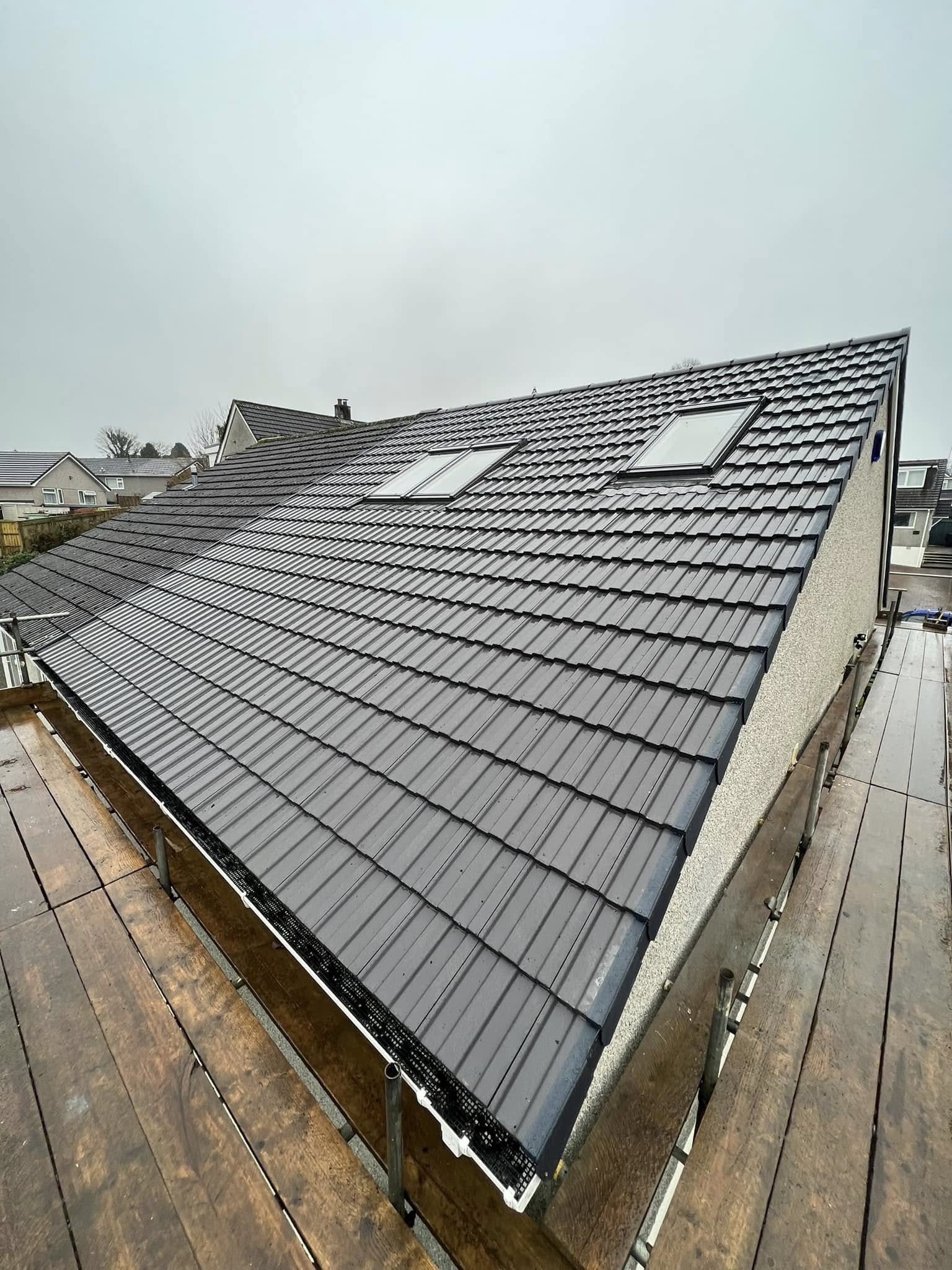 A view of a sloped roof with clean, dark tiles and two skylights, surrounded by scaffolding on a cloudy day, overlooking neighboring houses.