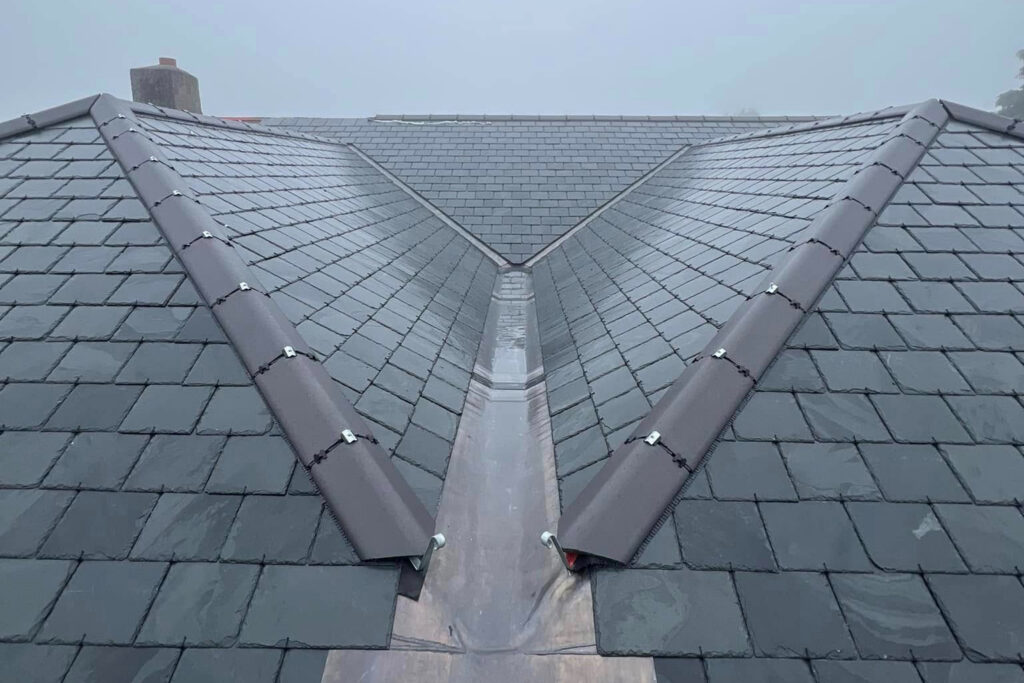 A close-up view of a slate roof with a central valley and capped ridges, taken on a foggy day. The tiles are dark gray, and the valley is lined with metal flashing.