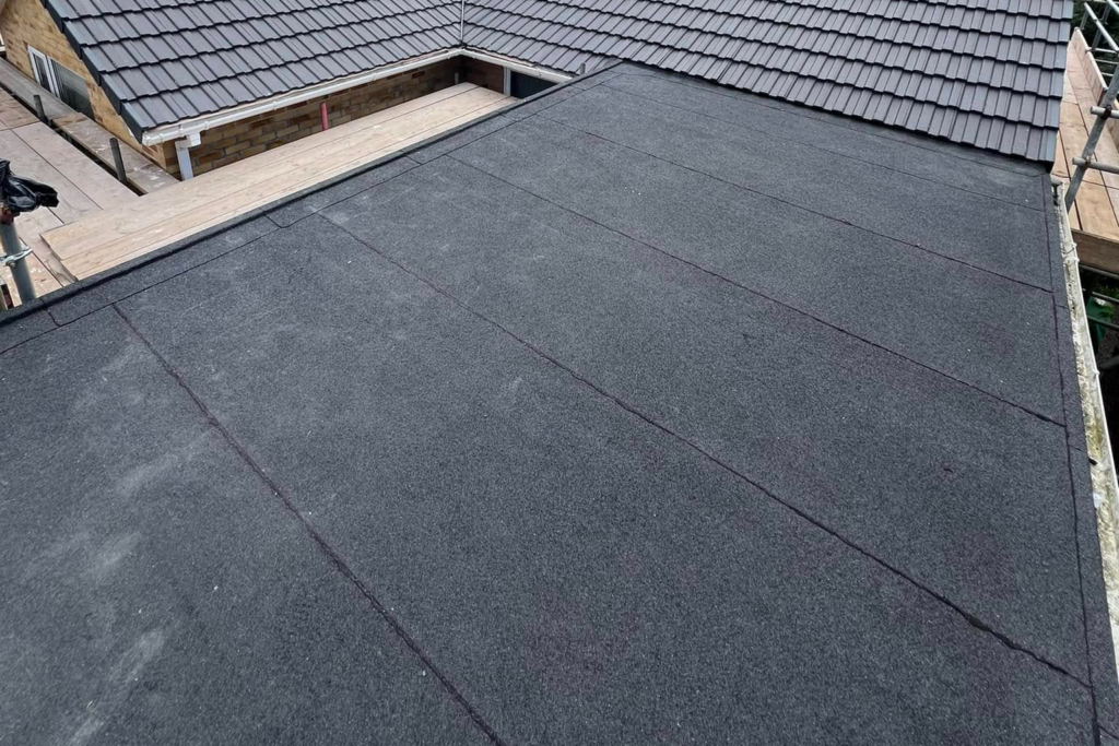 Flat roof covered with dark rubber or asphalt roofing material, featuring visible seam lines. In the background, a sloped roof with gray shingles and part of a wooden structure are visible.