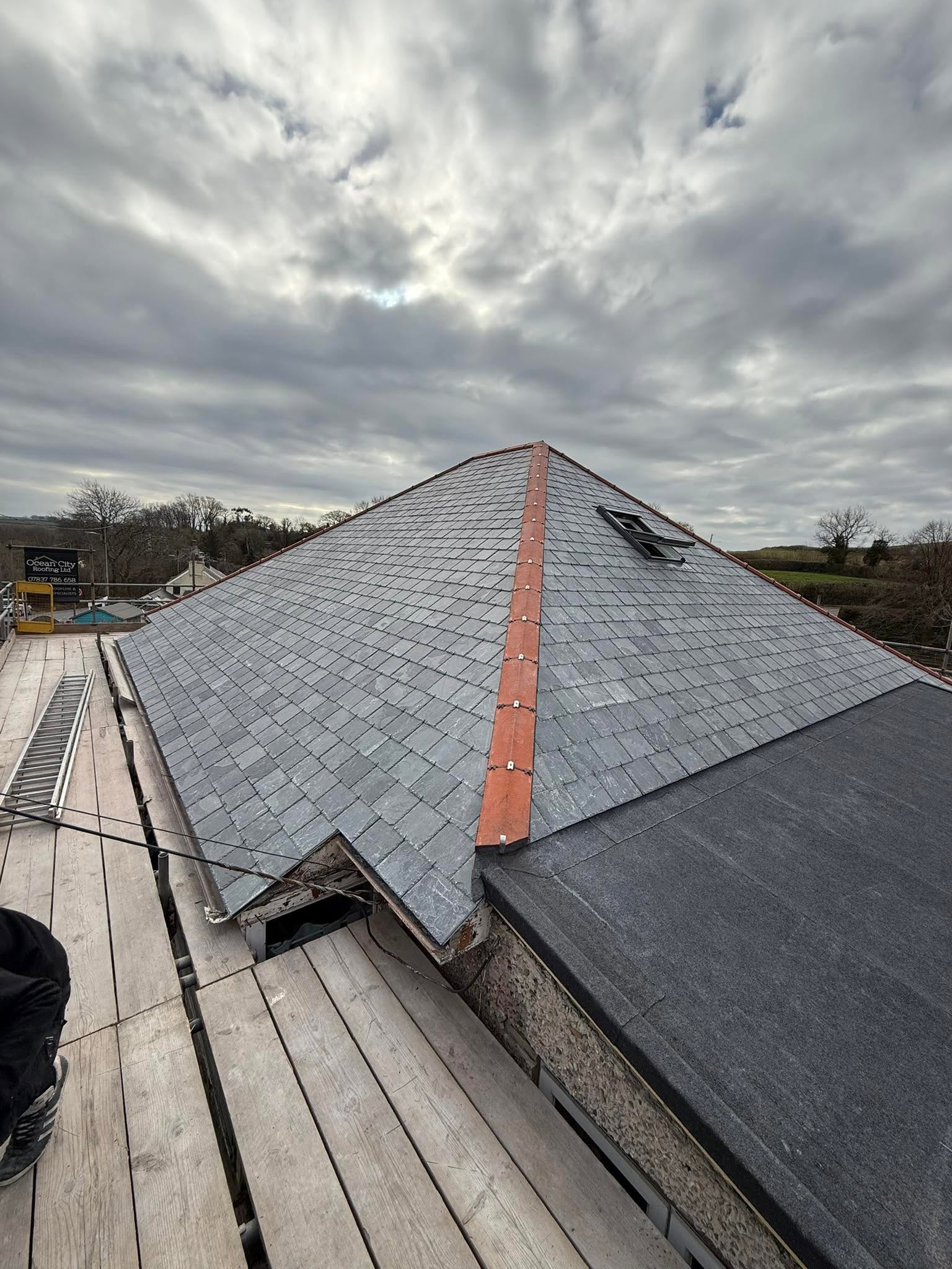 A sloped slate roof with a red ridge cap, small skylight, and cloudy sky above. Scaffolding surrounds the roof, and trees and fields are visible in the background.