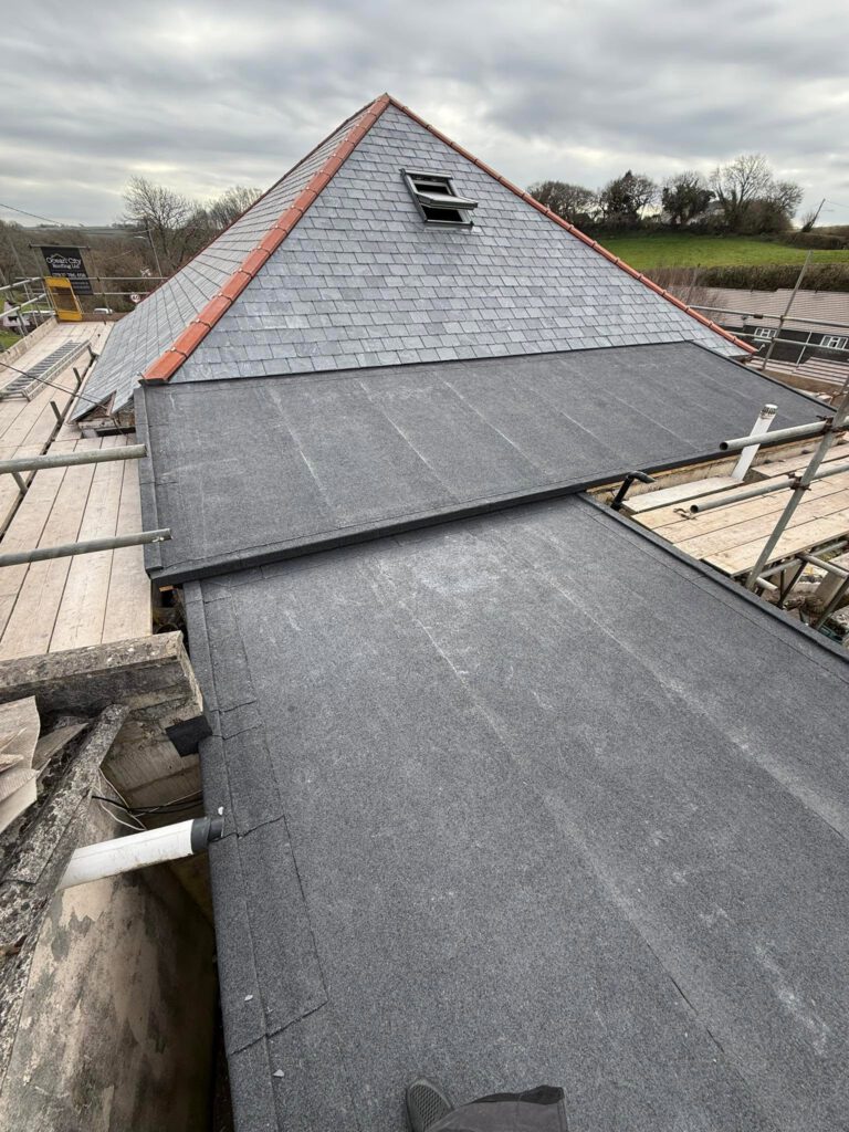 A view of a pitched gray slate roof with a small skylight, next to a flat black roof section, surrounded by scaffolding and cloudy skies.