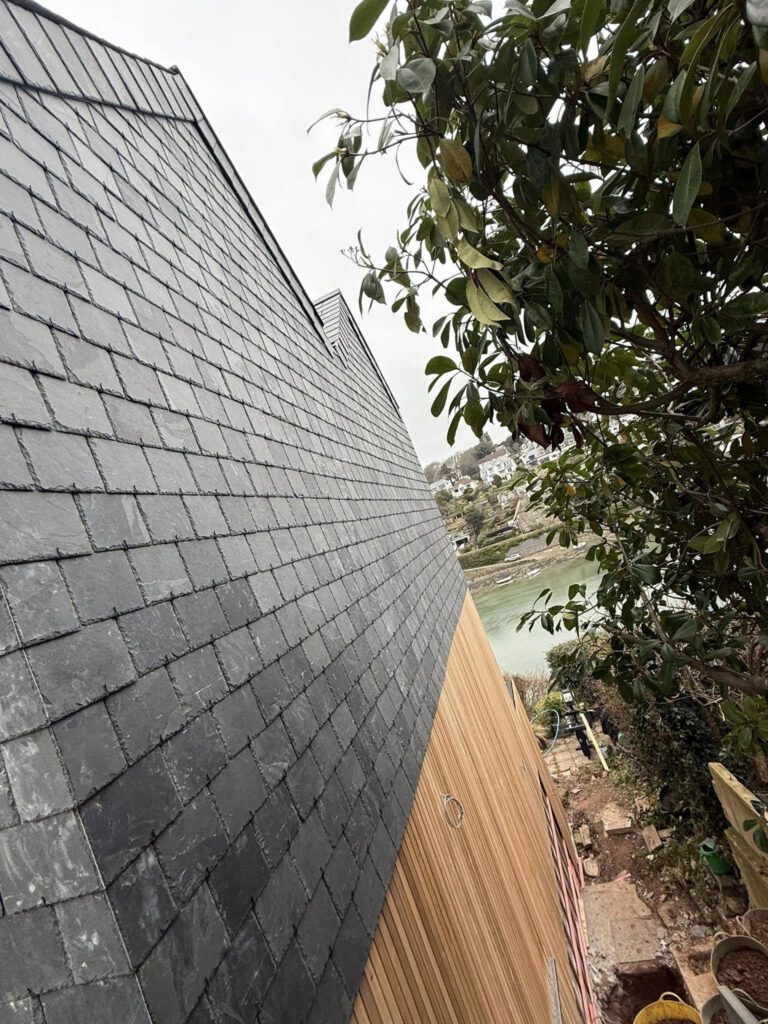 A slate-tiled sloped roof beside a wooden wall, with a leafy tree to the right and a river and houses visible in the background under a cloudy sky.