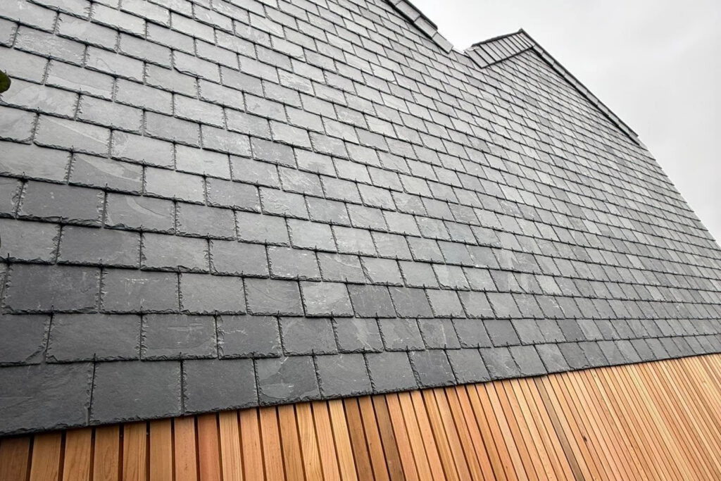 Close-up view of a building exterior featuring dark gray slate tiles above vertical wooden slats, shot from a low angle against a cloudy sky.