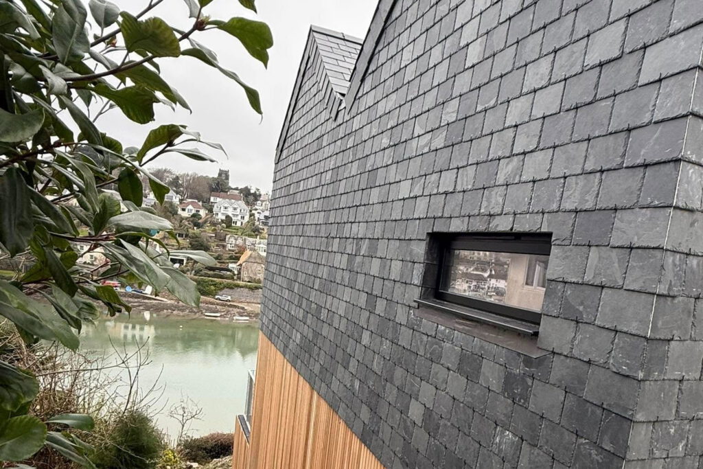 A close-up of a house with dark gray slate shingles and a small window, overlooking a river and a village with houses in the background. Green leaves frame part of the image on the left.