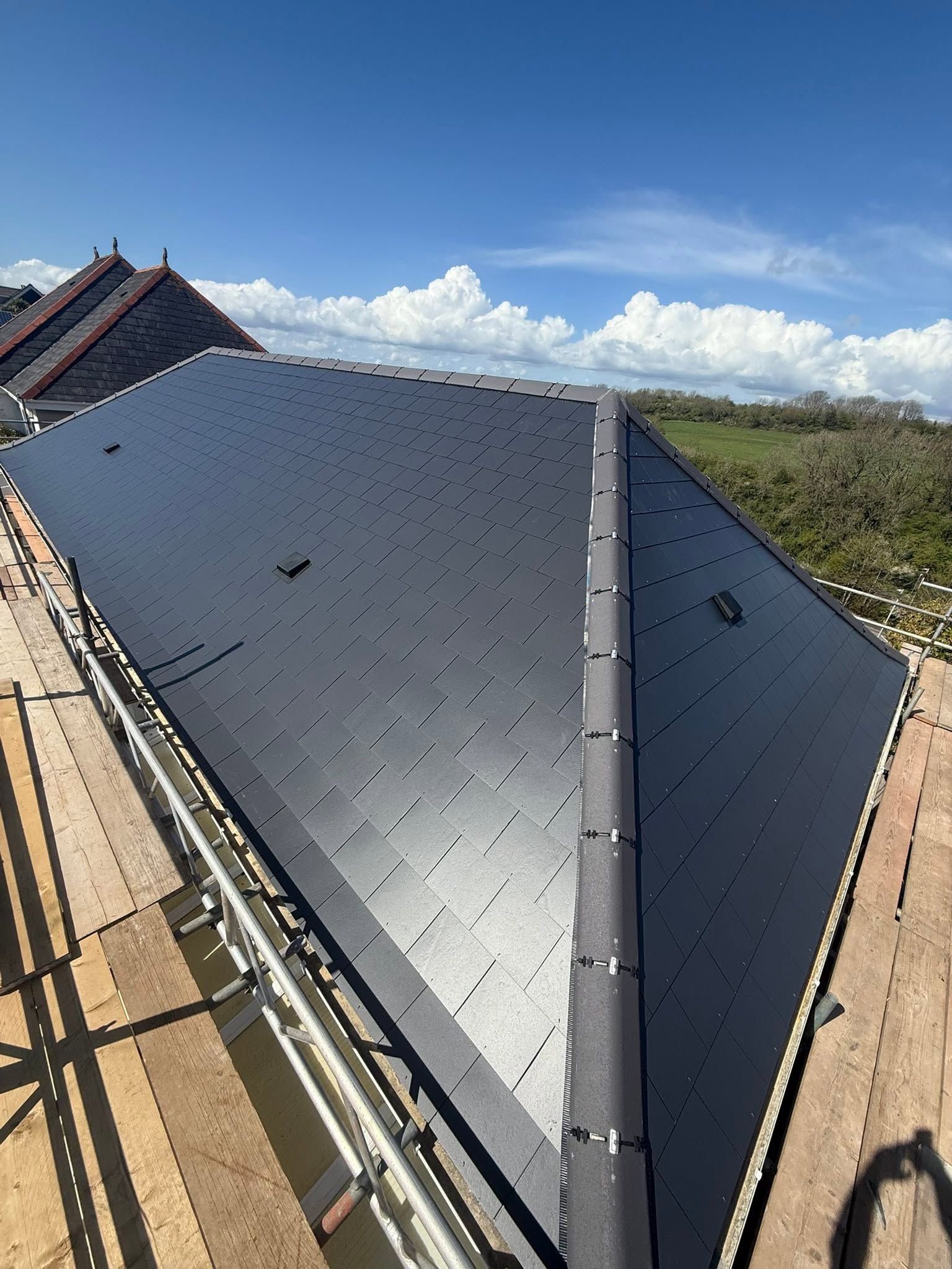 A newly constructed slate roof with clean, dark shingles under a bright blue sky, bordered by scaffolding and overlooking a green rural landscape.
