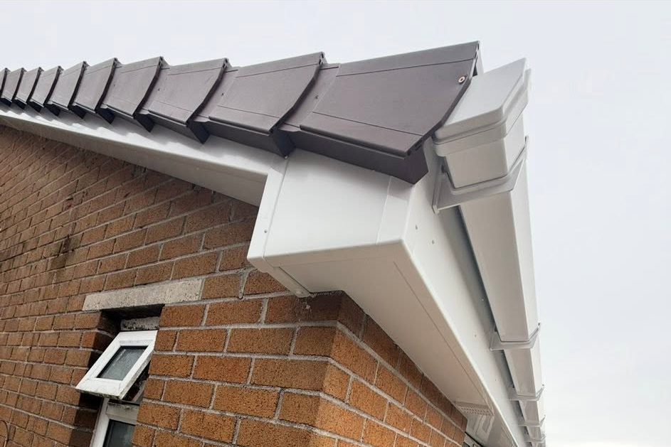 Close-up view of a house’s roof edge showing brown roof tiles, white plastic fascia, soffit boards, and guttering, with a small window on the brick wall below.