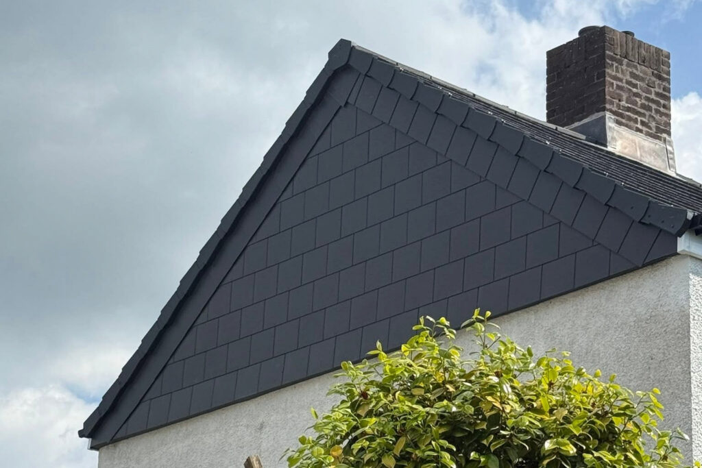 A close-up of a house roof with dark gray shingles, a brick chimney, white exterior walls, and a leafy green bush in the foreground under a cloudy sky.
