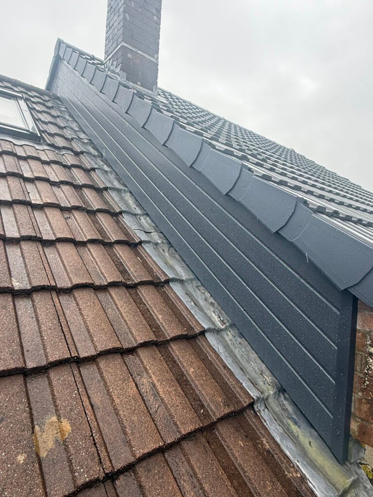 A close-up view of a rooftop shows brown roof tiles next to a dark grey metal cladding section, with a brick chimney and cloudy sky in the background.
