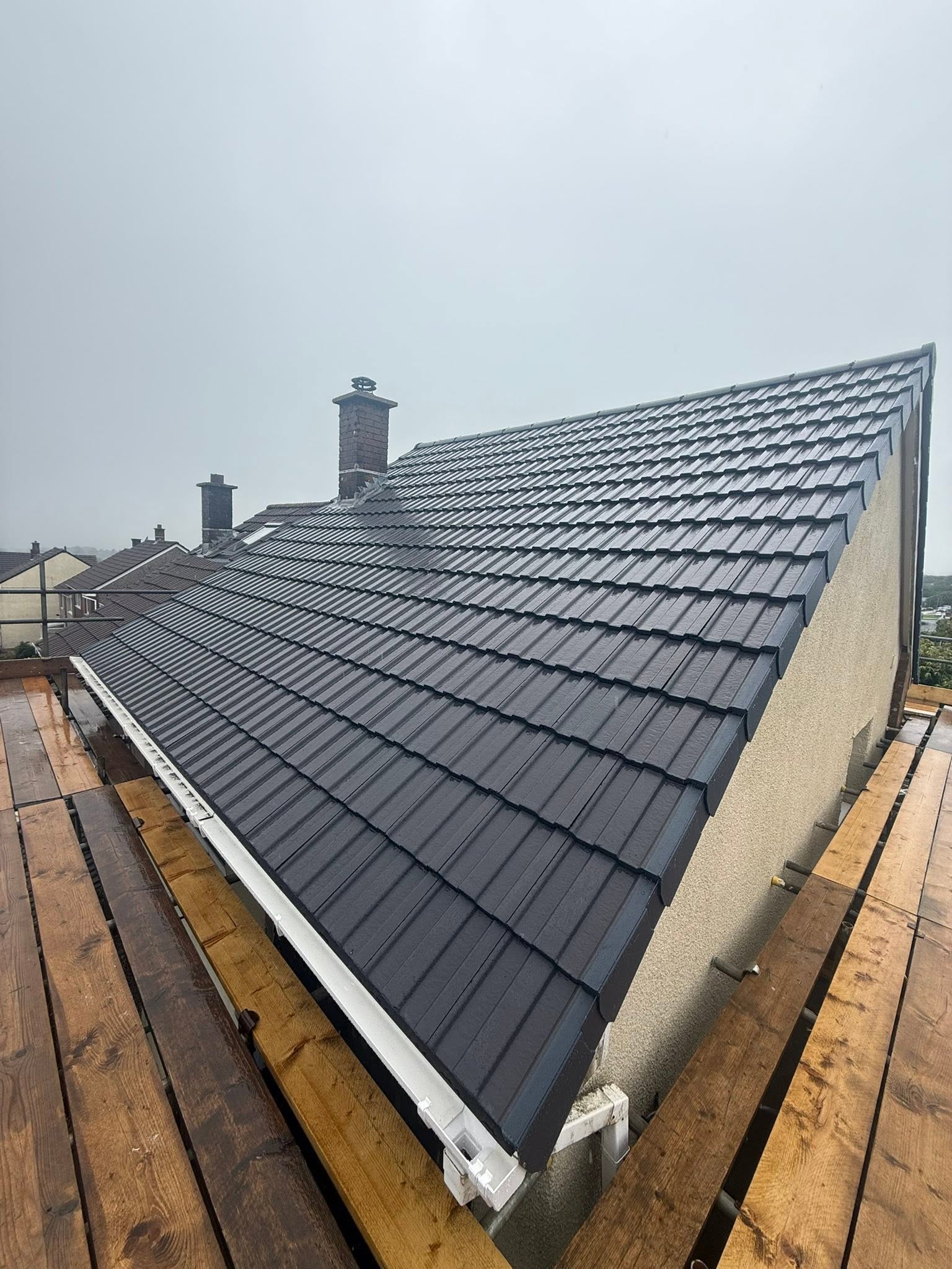 A sloped roof with dark gray tiles on a house, surrounded by wooden scaffolding. The sky is overcast, and neighboring rooftops are visible in the background.
