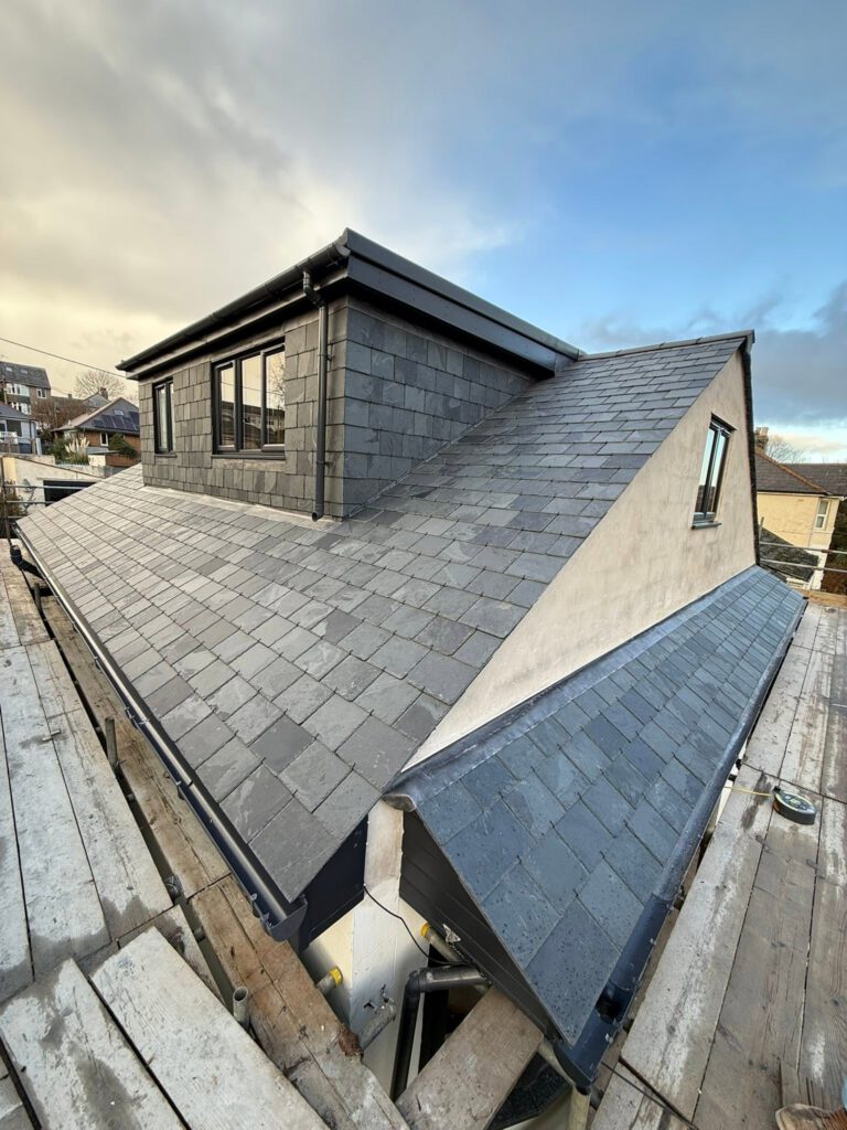 A house with a newly constructed slate roof and dormer window, surrounded by scaffolding under a partly cloudy sky.