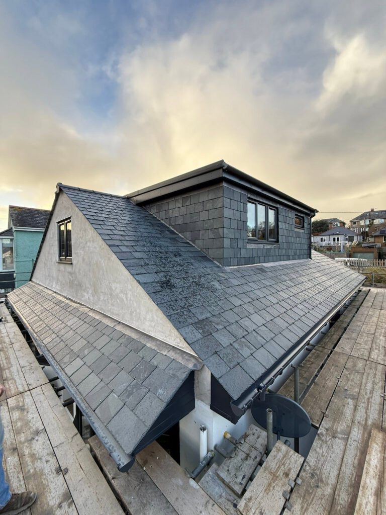 A modern house roof with grey slate tiles and dormer windows, surrounded by scaffolding under a partly cloudy sky. Nearby houses are visible in the background.