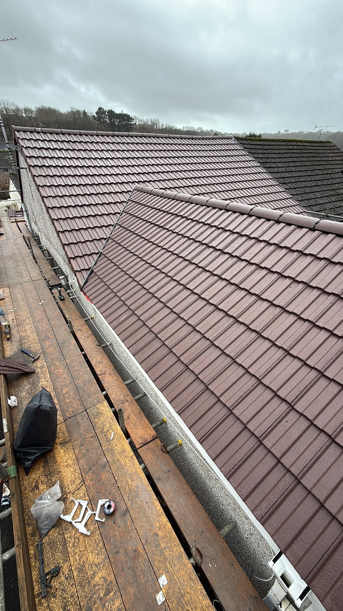 A view from scaffolding shows a freshly tiled roof on a house, with tools and materials scattered on the wooden platform. The sky is overcast.