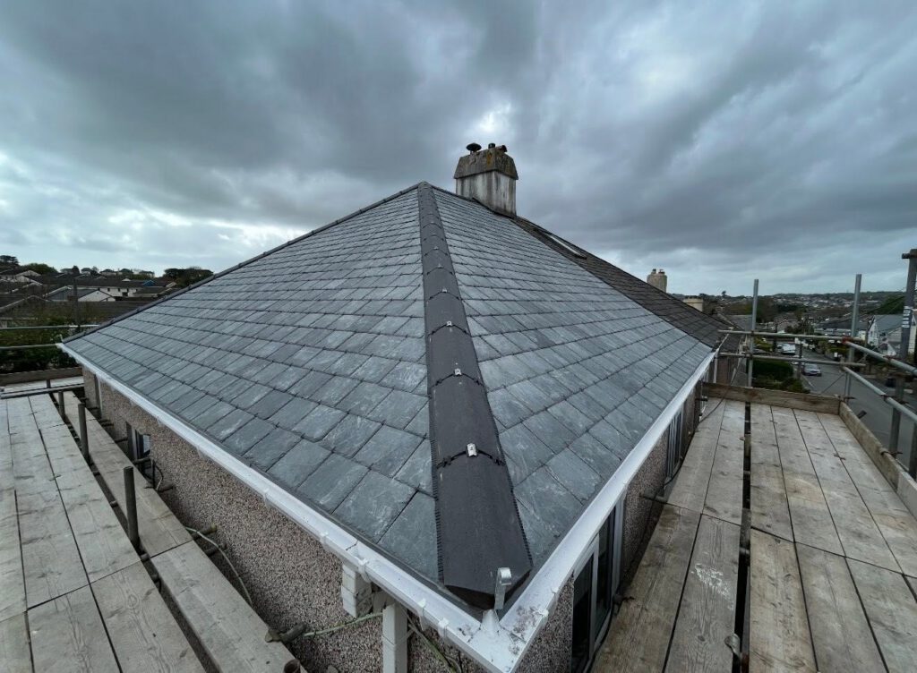 A pitched slate roof with dark ridge caps, surrounded by scaffolding, under a cloudy sky in a residential area.