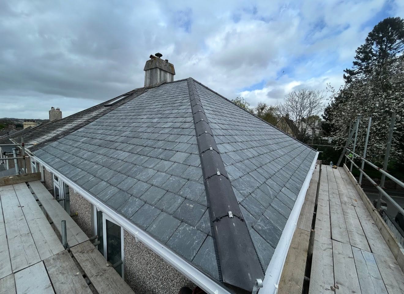 A view of a gray slate tiled roof with a central ridge, scaffolding on both sides, and cloudy sky above. Trees and neighboring houses are visible in the background.