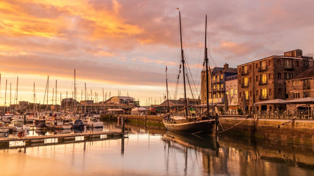 Sailboats docked in a calm harbor at sunset, with historic brick buildings and glowing sky reflecting on the water.