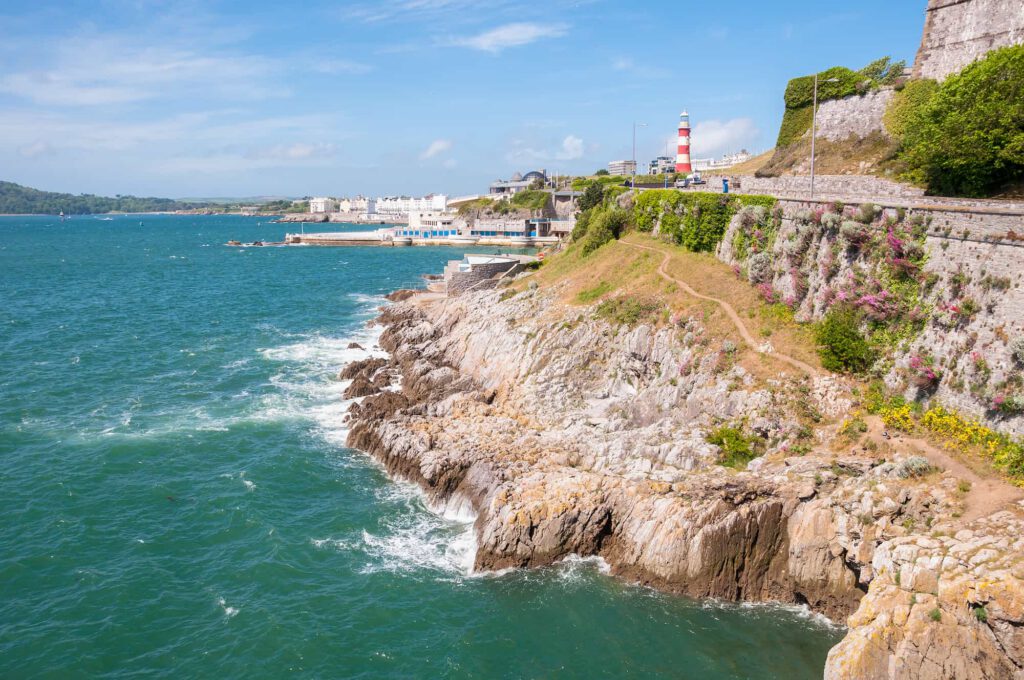 Rocky coastline with waves, a winding path along the cliff, and a red-and-white lighthouse in the distance under a blue sky with scattered clouds.