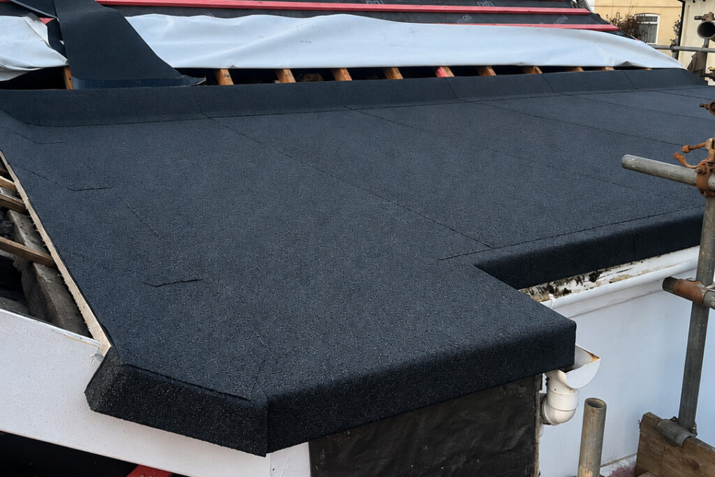 A close-up of a roof under construction, featuring newly installed dark roofing felt and visible wooden beams beneath an unfinished upper layer. Scaffolding and construction materials surround the structure.