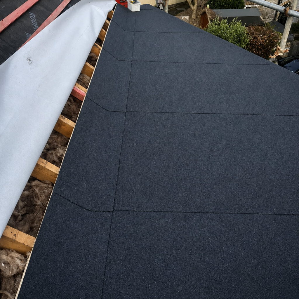 Close-up view of a roof being constructed, featuring black asphalt shingles partially installed, wooden beams, and exposed insulation beneath a section of protective underlayment.